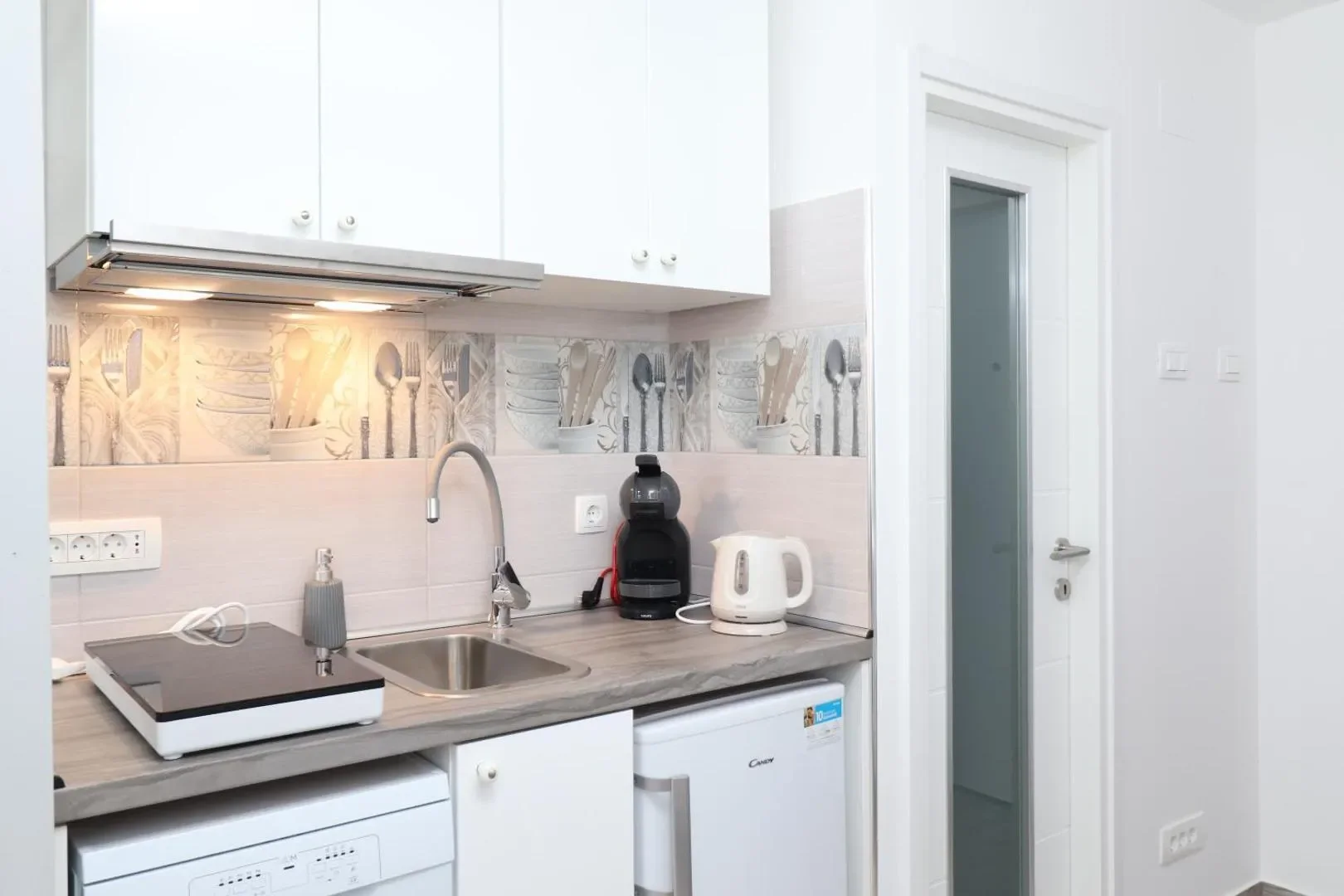 Kitchen counter with a sink, cooktop, kettle and coffee machine beneath white cupboards.